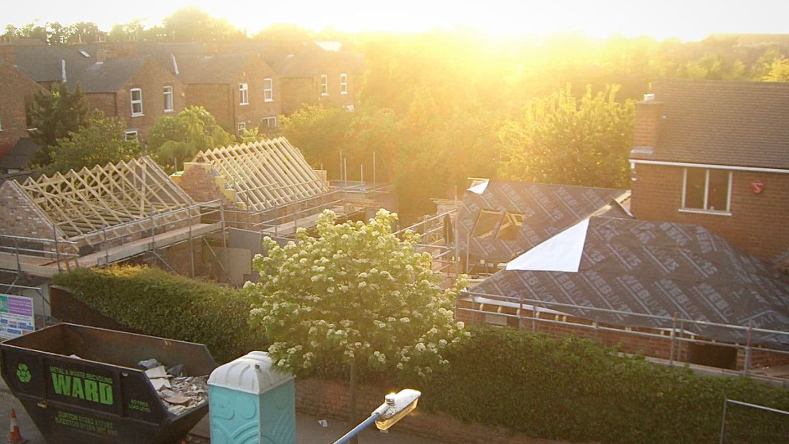 A build in progress at sunset: new timber roof trusses and felted roofs going on, with scaffolding and a skip in the foreground.