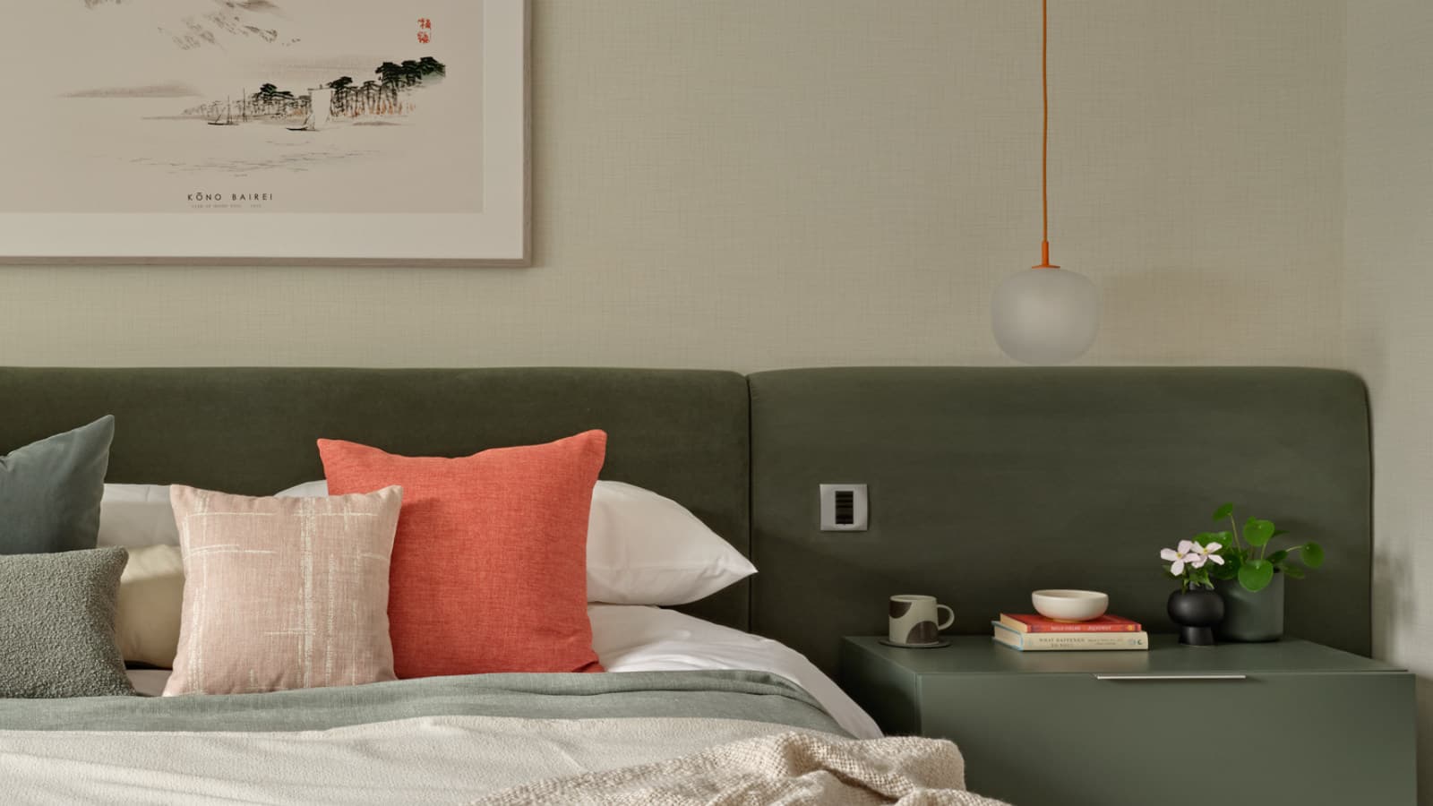 A bed with a khaki green fitted headboard, a pendant bedside light, green bedside table and framed artwork above.