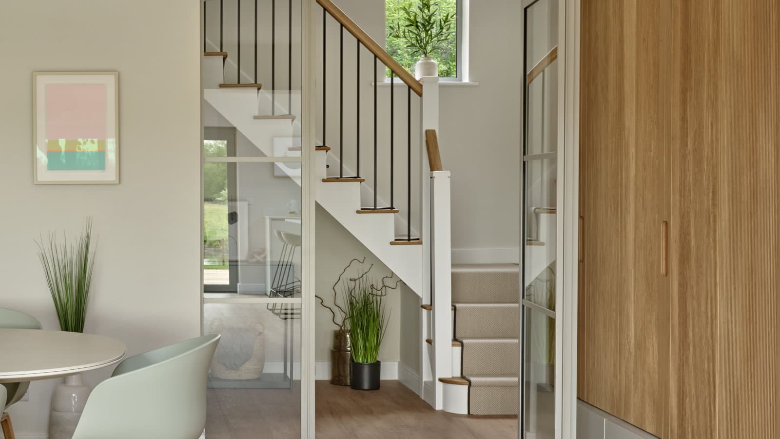 A staircase with a white balustrade, black spindles and oak handrail, beside tall fitted cupboard doors and a dining table.