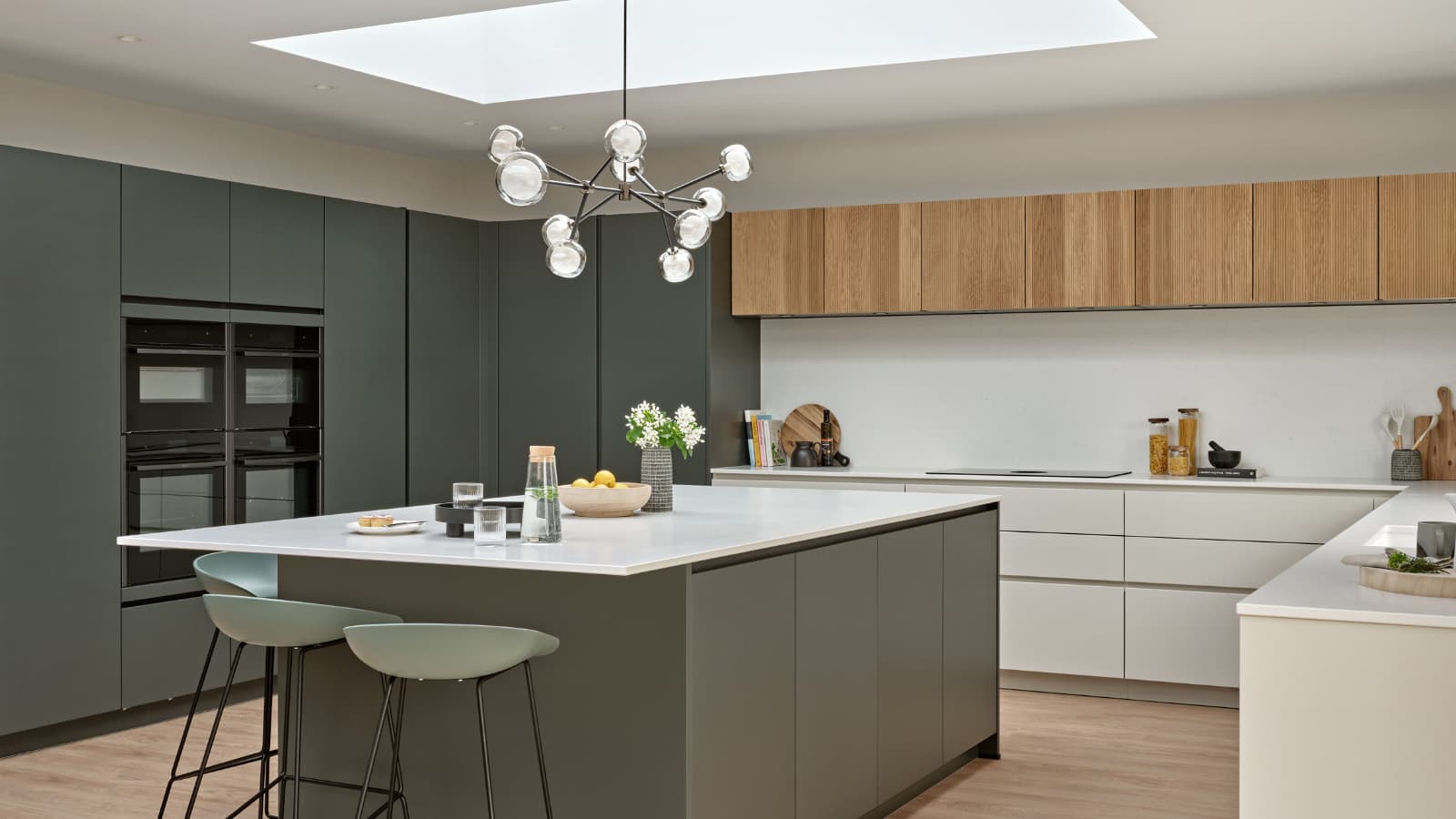 A modern open-plan kitchen with grey-green handleless cabinets, wood-fronted wall units and a white island with pale green bar stools, lit by a large skylight and a glass-globe pendant.
