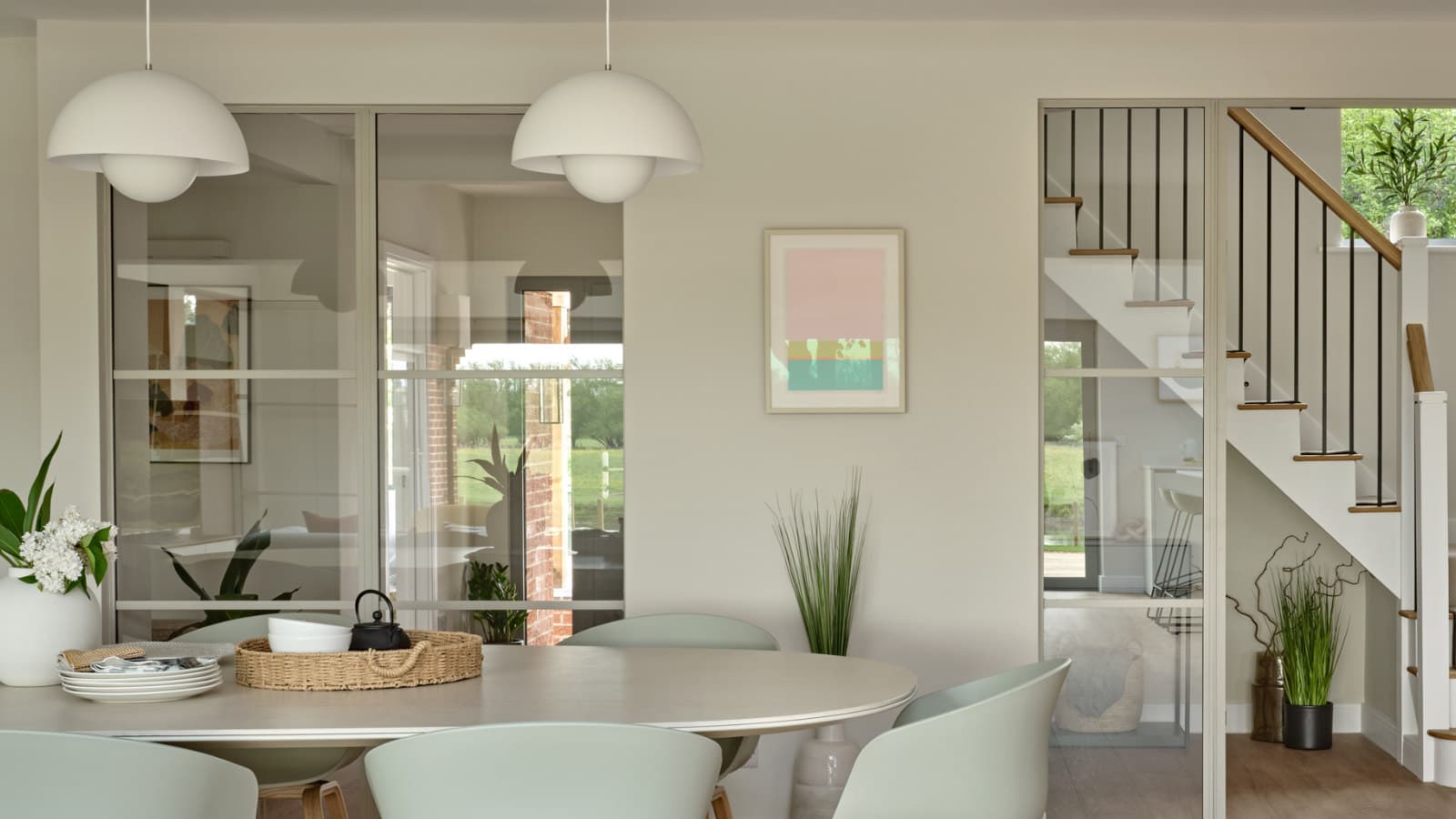 Dining room with two white pendant lights over a pale oval table, a slim-framed internal glazed screen behind it, and a second glazed screen to the right looking through to the staircase and garden