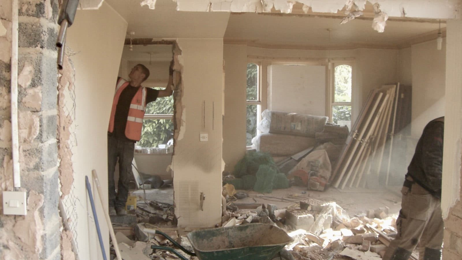 A house interior mid-demolition, two workers breaking out an internal wall, one in an orange hi-vis vest, dust hanging in the light and a wheelbarrow amid broken masonry on the floor.
