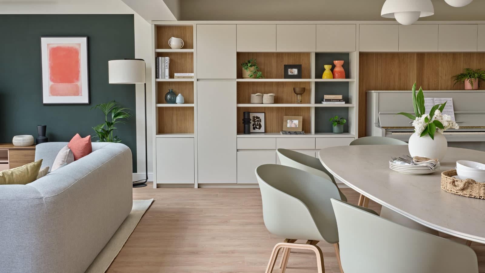 An open-plan living and dining room with floor-to-ceiling fitted joinery: cream cabinets framing oak-lined open display shelves, beside a dining table, sofa and upright piano.